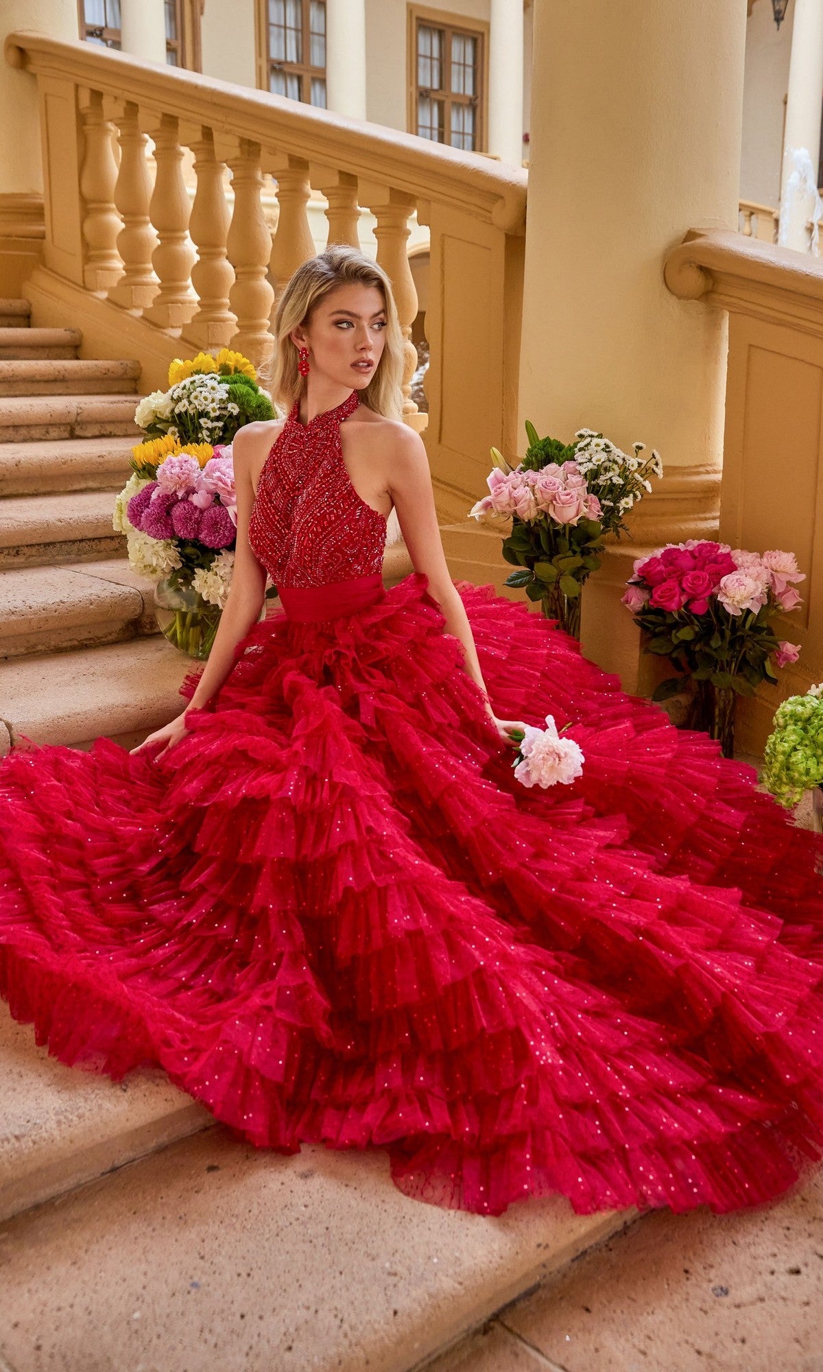 A woman wearing the Ava Presley 47258 long prom dress poses on stone steps with a small bouquet, surrounded by vibrant flower arrangements and beige railings.